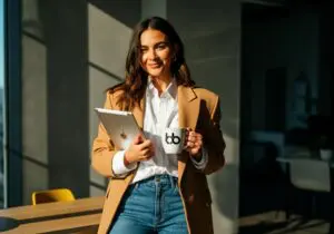 Woman in a camel blazer holding a tablet and branded mug in a sunlit modern office, smiling with confident editorial poise.