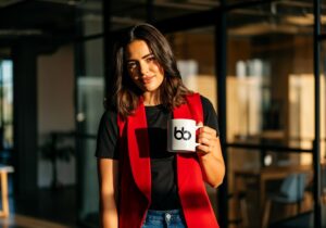 Professional in a bold red vest and black tee holding a branded mug near glass-walled meeting rooms, exuding confident, fashion-forward style, get featured in Vogue.