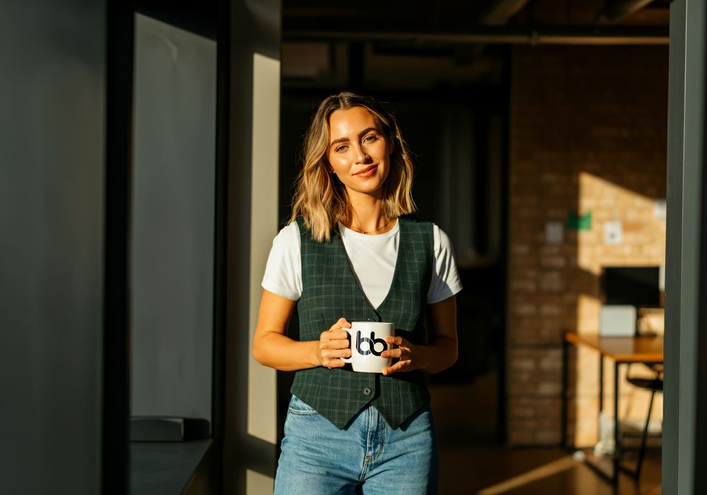 Woman in a green plaid vest and white tee holding a branded mug in warm afternoon light inside a modern office, projecting chic editorial poise, get featured in Vogue.